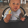 baby, child, high_chair, eating, snack, food, hand, smiling, pouting, portrait, indoor, dining_area, chair, table, wood_floor, home, gray_shirt, curly_hair, toddler, photography