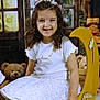 child, girl, smiling, curly_hair, white_skirt, white_shirt, headband, flower_crown, yellow_crescent, decorative, teddy_bear, plush_toy, window_backdrop, rustic, indoor, portrait, happy, seated, white_socks, gold_shoes