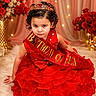 child, toddler, birthday, crown, dress, red_dress, sash, birthday_queen, portrait, sitting, curly_hair, smiling, indoor, floral_decor, roses, fairy_lights, celebration, phone_screenshot, royal_theme, photography