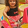 child, toddler, curly_hair, polka_dot, dress, toy_car, indoor, play_area, colorful, cartoon, smiling, fun, cute, seat, vehicle, handle, wheel, happy, face, person