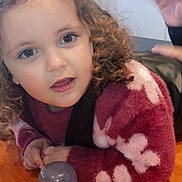 Noëlia a rejoint le concours — aidez-le/la à gagner de superbes lots ! child, curly_hair, sweater, maroon, white_flowers, table, wooden_table, cup, transparent_cup, hand, face, eyes, indoor, playful, young_child, person, cozy, smiling, closeup, curious