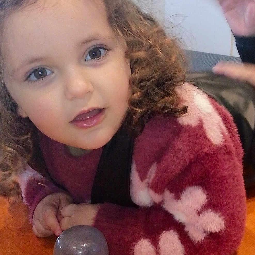 Noëlia a rejoint le concours — aidez-le/la à gagner de superbes lots ! child, closeup, cozy, cup, curious, curly_hair, eyes, face, hand, indoor, maroon, person, playful, smiling, sweater, table, transparent_cup, white_flowers, wooden_table, young_child