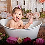 baby, toddler, bathtub, white_tub, flowers, pink_bow, blue_eyes, splash, water_droplets, smile, surprised, studio_backdrop, wooden_floor, portrait, skin, happy, playful, indoors, rosy_cheeks, prop_decor