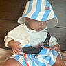 baby, child, hat, bucket_hat, striped_clothing, shorts, white_shirt, sitting, indoor, wooden_background, blanket, cute, chubby_legs, curious, young_child, portrait, casual_clothing, infant, soft_texture, looking_down