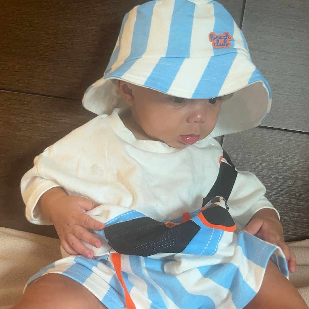 Andrei is registered to the contest to win money with this photo: baby, child, hat, bucket_hat, striped_clothing, shorts, white_shirt, sitting, indoor, wooden_background, blanket, cute, chubby_legs, curious, young_child, portrait, casual_clothing, infant, soft_texture, looking_down