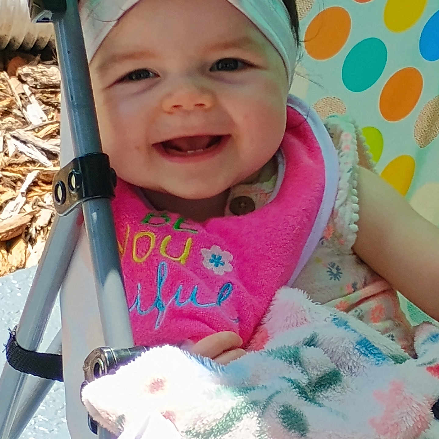 Aubree is registered to the contest to win money with this photo: baby, child, smiling, headband, pink_bib, stroller, blanket, colorful_background, happy, face, infant, outdoor, clothing, person, portrait, cute, joyful, seat, baby_girl, playful