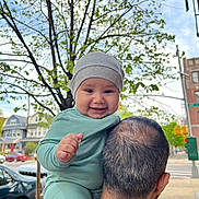 Ismail is registered to the contest to win money with this photo: adult, baby, car, child, daylight, face, green_clothing, happy, hat, head, house, leaves, outdoor, person, portrait, shoulder, smiling, street, tree, urban