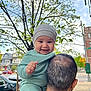adult, baby, car, child, daylight, face, green_clothing, happy, hat, head, house, leaves, outdoor, person, portrait, shoulder, smiling, street, tree, urban