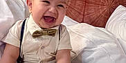 Ismail joined the competition — help win amazing prizes! baby, bed, bow_tie, child, clothing, cute, feet, hands, happy, headboard, indoors, infant, joyful, person, portrait, sitting, skin, smiling, suspenders, white_bed