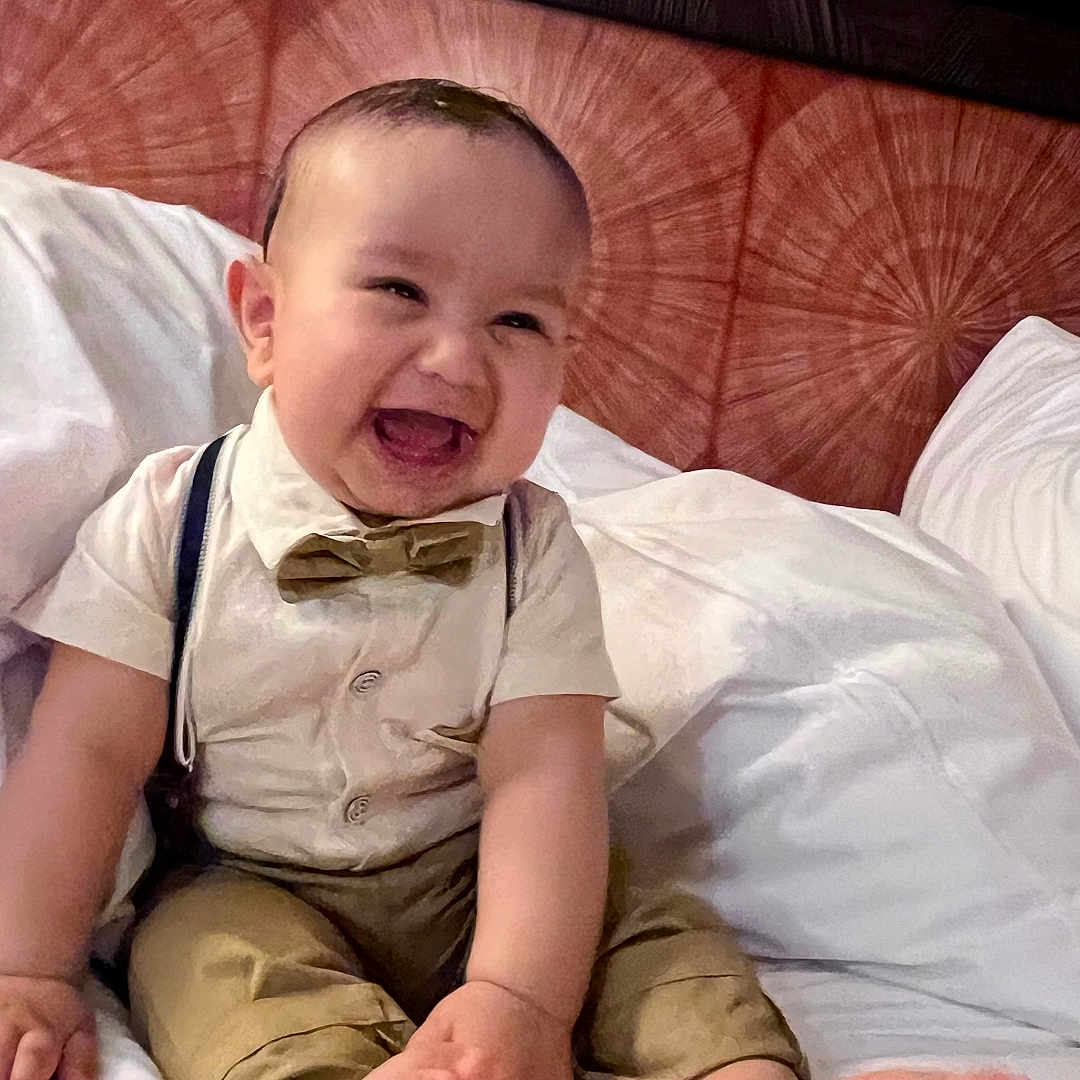 Ismail joined the competition — help win amazing prizes! baby, bed, bow_tie, child, clothing, cute, feet, hands, happy, headboard, indoors, infant, joyful, person, portrait, sitting, skin, smiling, suspenders, white_bed