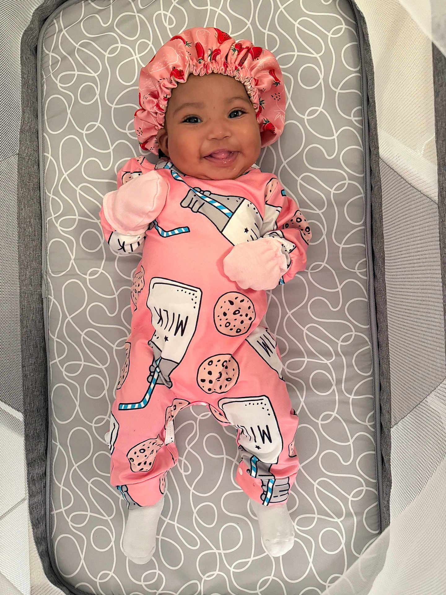 Genesis is registered to the contest to win money with this photo: baby, infant, smiling, pink_onesie, bonnet, milk_print, cookie_print, crib, mattress, patterned_bedding, happy, tongue_out, lying_down, cute, child, soft_lighting, indoors, cozy, comfortable, playpen