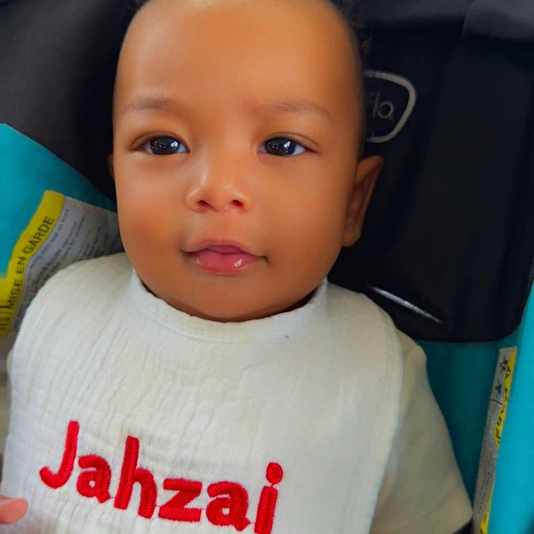 Jahzai is registered to the contest to win money with this photo: baby, child, bib, stroller, face, portrait, cute, infant, clothing, smile, young_child, seated, indoors, person, head, skin, expression, baby_bib, fabric, closeup