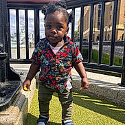 Ikraam is registered to the contest to win money with this photo: toddler, child, portrait, outdoor, sunny, grass, urban, railing, building, pigtails, shirt, sneakers, curious, standing, daylight, person, fashion, casual, young_child, expression