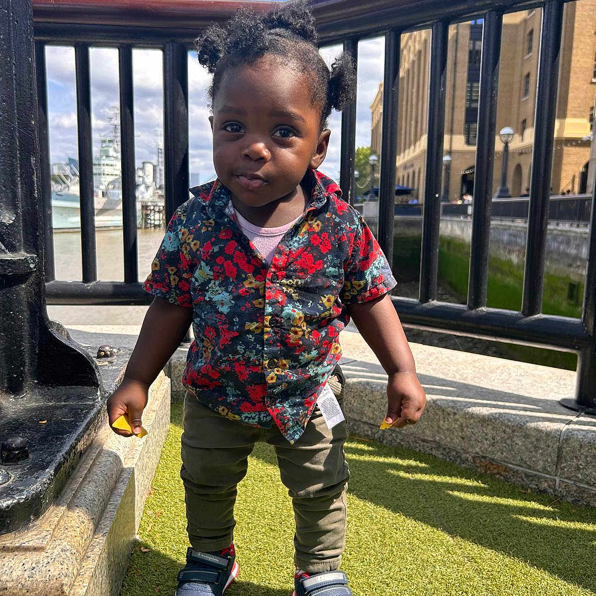 Ikraam is registered to the contest to win money with this photo: building, casual, child, curious, daylight, expression, fashion, grass, outdoor, person, pigtails, portrait, railing, shirt, sneakers, standing, sunny, toddler, urban, young_child