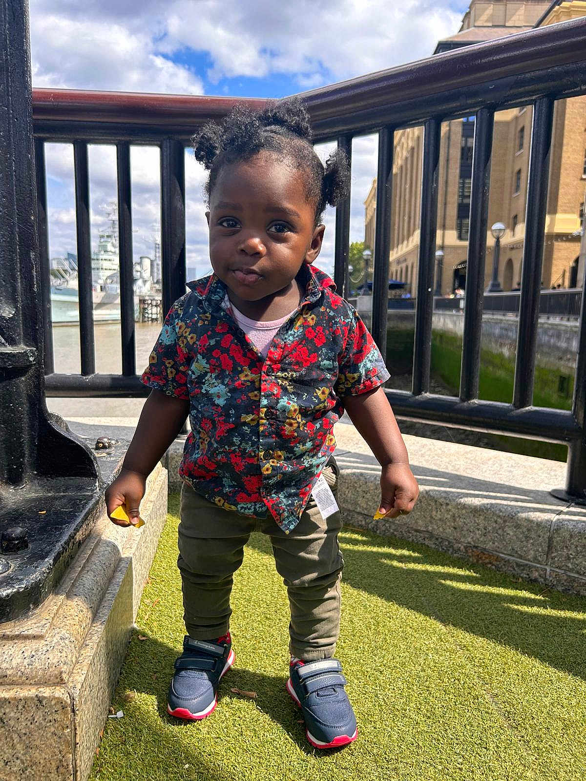 Ikraam is registered to the contest to win money with this photo: toddler, child, portrait, outdoor, sunny, grass, urban, railing, building, pigtails, shirt, sneakers, curious, standing, daylight, person, fashion, casual, young_child, expression