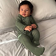 Khalil is registered to the contest to win money with this photo: baby, infant, green_onesie, bed, pillow, white_bed_sheet, face, hands, feet, sleepwear, child, young, portrait, indoors, natural_light, resting, cute, comfortable, smiling, lying_down