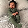 baby, infant, green_onesie, bed, pillow, white_bed_sheet, face, hands, feet, sleepwear, child, young, portrait, indoors, natural_light, resting, cute, comfortable, smiling, lying_down