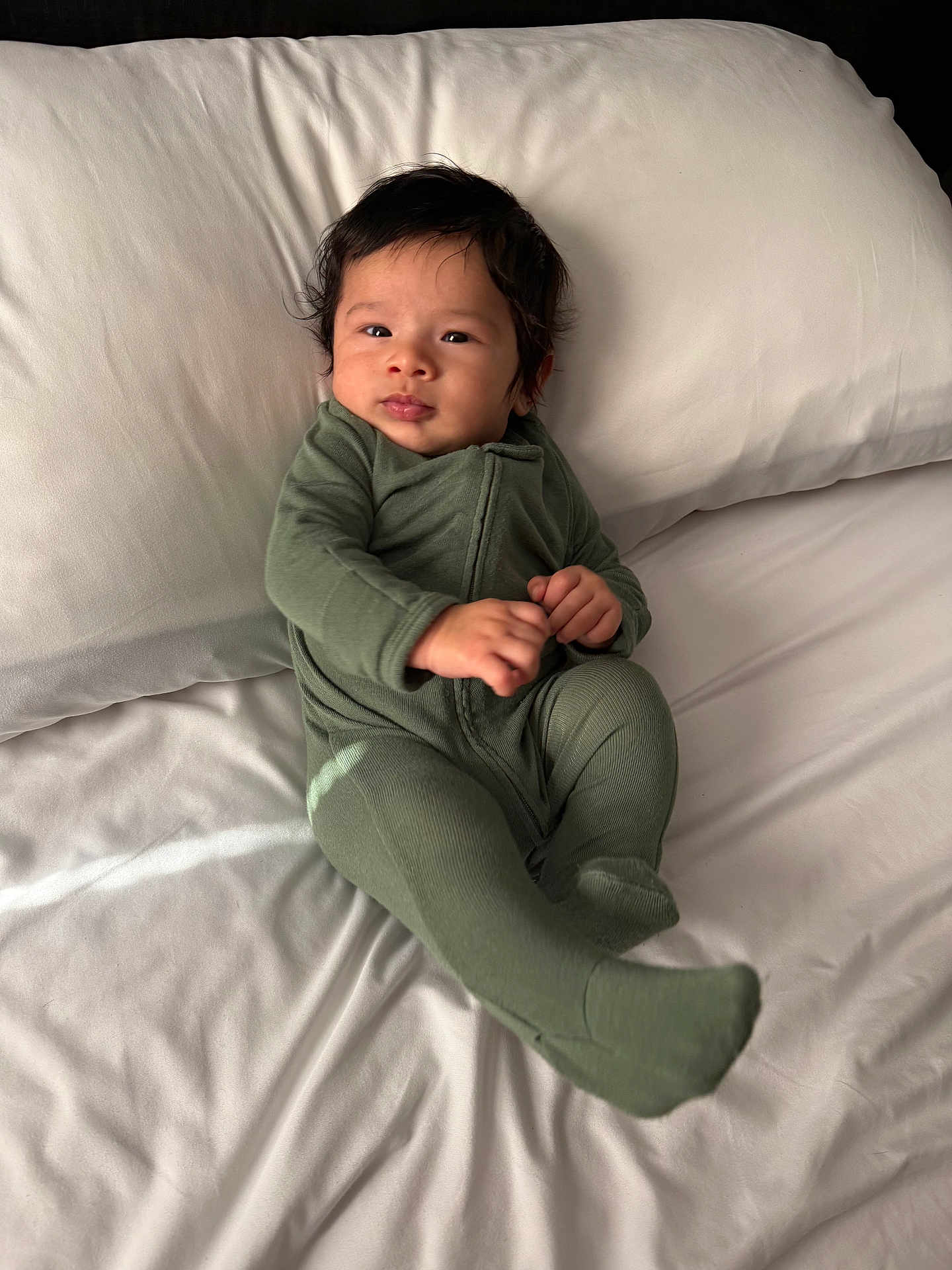 Khalil is registered to the contest to win money with this photo: baby, infant, green_onesie, bed, pillow, white_bed_sheet, face, hands, feet, sleepwear, child, young, portrait, indoors, natural_light, resting, cute, comfortable, smiling, lying_down