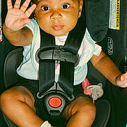 Isla is registered to the contest to win money with this photo: baby, child, car_seat, safety_harness, white_bow, sock, pacifier, infant, cute, waving_hand, seat_belt, portrait, indoor, young_child, black_clothing, baby_clothing, face, looking_at_camera, sitting, adorable