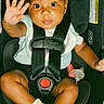 baby, child, car_seat, safety_harness, white_bow, sock, pacifier, infant, cute, waving_hand, seat_belt, portrait, indoor, young_child, black_clothing, baby_clothing, face, looking_at_camera, sitting, adorable