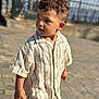 child, boy, curly_hair, striped_shirt, short_sleeves, pavement, outdoor, daylight, portrait, standing, casual_clothing, background_blur, fence, curious, person, young_child, summer, sunlight, side_view, urban_outdoor
