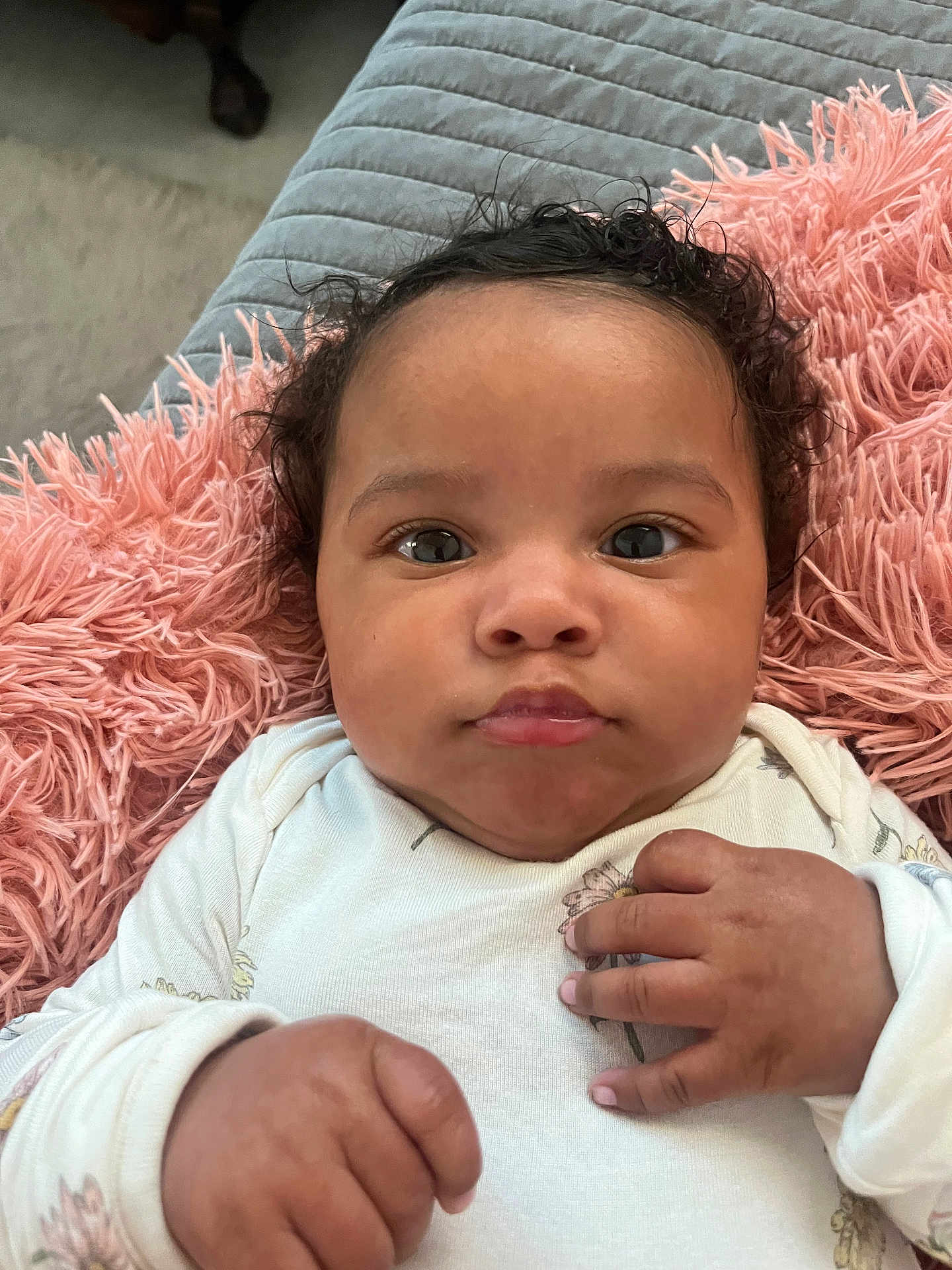 Isla joined the competition — help win amazing prizes! baby, infant, close_up, face, hands, curly_hair, blanket, pink, quilted, onesie, floral, soft_texture, lying_down, indoors, portrait, cute, child, skin, expression, young