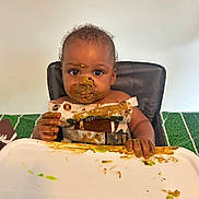 Tajhire is registered to the contest to win money with this photo: baby, brown_food, child, clothing, curiosity, curly_hair, eating, expression, face_paint, food, green_food, hands, high_chair, indoor, meal, messy, plate, smudged, table, toddler