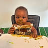 toddler, baby, high_chair, messy, food, face_paint, curly_hair, eating, green_food, brown_food, plate, table, clothing, child, indoor, expression, hands, smudged, meal, curiosity