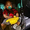 baby, pacifier, car_seat, plush_toy, child, infant, seatbelt, red_shirt, gray_shorts, sock, indoors, vehicle_interior, black_background, sitting, toy, child_safety, cute, young_child, comfort, car