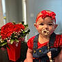 baby, infant, pacifier, headband, bow, red_clothing, denim_overalls, bouquet, flowers, vase, sitting, portrait, indoor, table, face_paint, pacifier_clip, bokeh, hands, heart_pattern, closeup