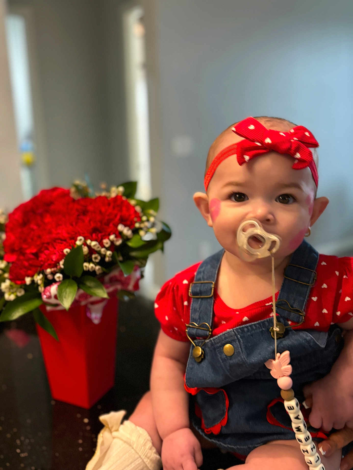 Ellie is registered to the contest to win money with this photo: baby, infant, pacifier, headband, bow, red_clothing, denim_overalls, bouquet, flowers, vase, sitting, portrait, indoor, table, face_paint, pacifier_clip, bokeh, hands, heart_pattern, closeup