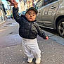 child, toddler, cap, puffer_jacket, sweatpants, shoes, sidewalk, street, urban, car, building, hand, holding_hands, city, portrait, outdoor, walking, neutral_expression, hat, cold_weather