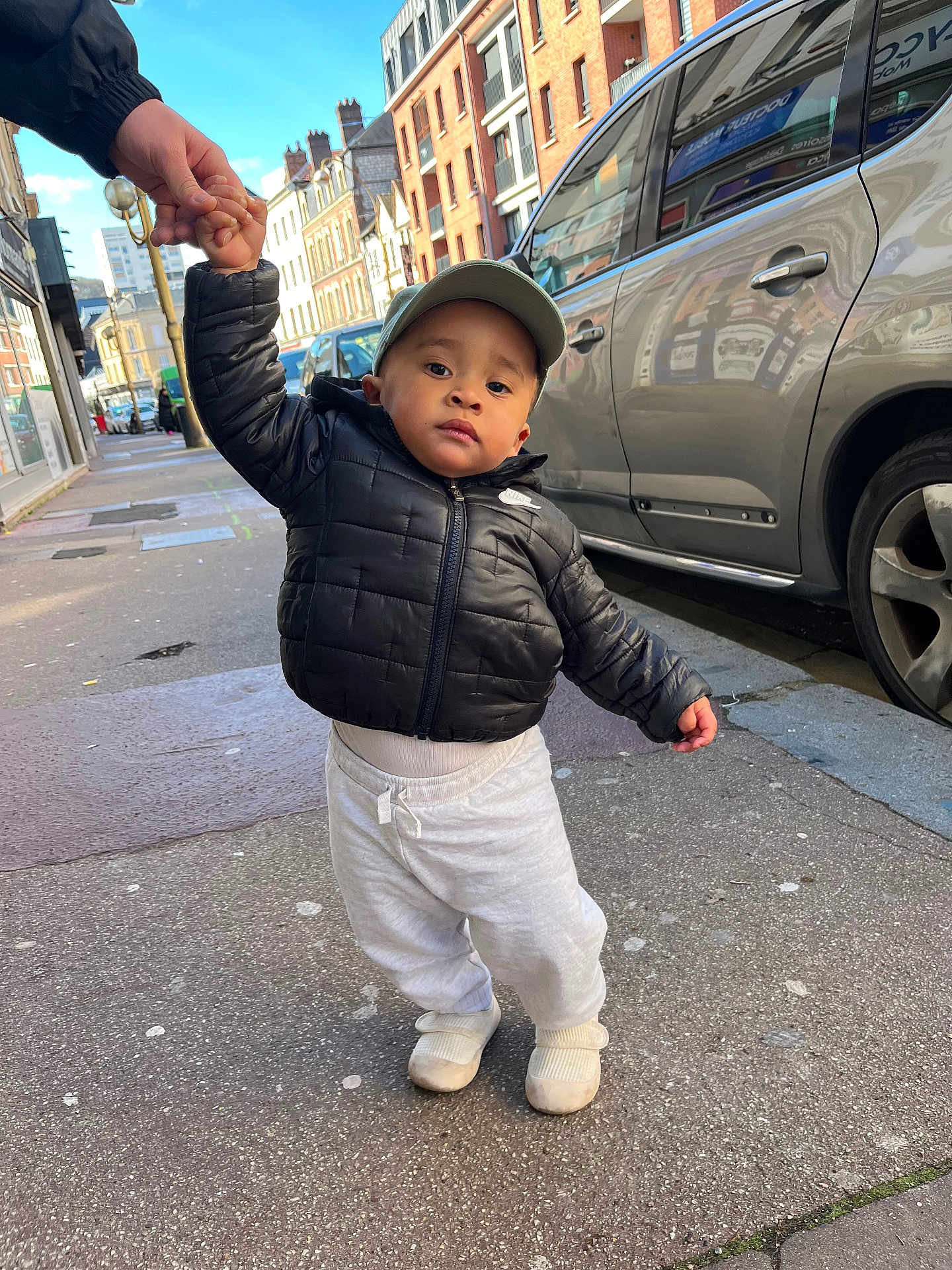 Issa participe au concours pour gagner de l'argent avec cette photo : child, toddler, cap, puffer_jacket, sweatpants, shoes, sidewalk, street, urban, car, building, hand, holding_hands, city, portrait, outdoor, walking, neutral_expression, hat, cold_weather