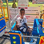 Naël participe au concours pour gagner de l'argent avec cette photo : child, boy, train_ride, smiling, outdoor, daylight, playground, sign, warning_sign, safety, blue, yellow, seat, clothing, shorts, tshirt, sandals, bell, metal, fun