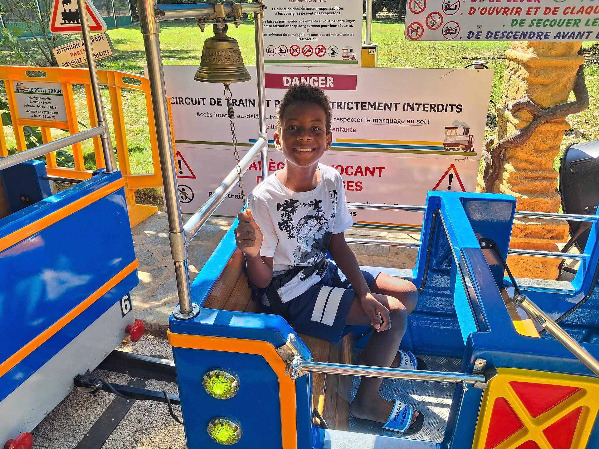 Naël participe au concours pour gagner de l'argent avec cette photo : child, boy, train_ride, smiling, outdoor, daylight, playground, sign, warning_sign, safety, blue, yellow, seat, clothing, shorts, tshirt, sandals, bell, metal, fun