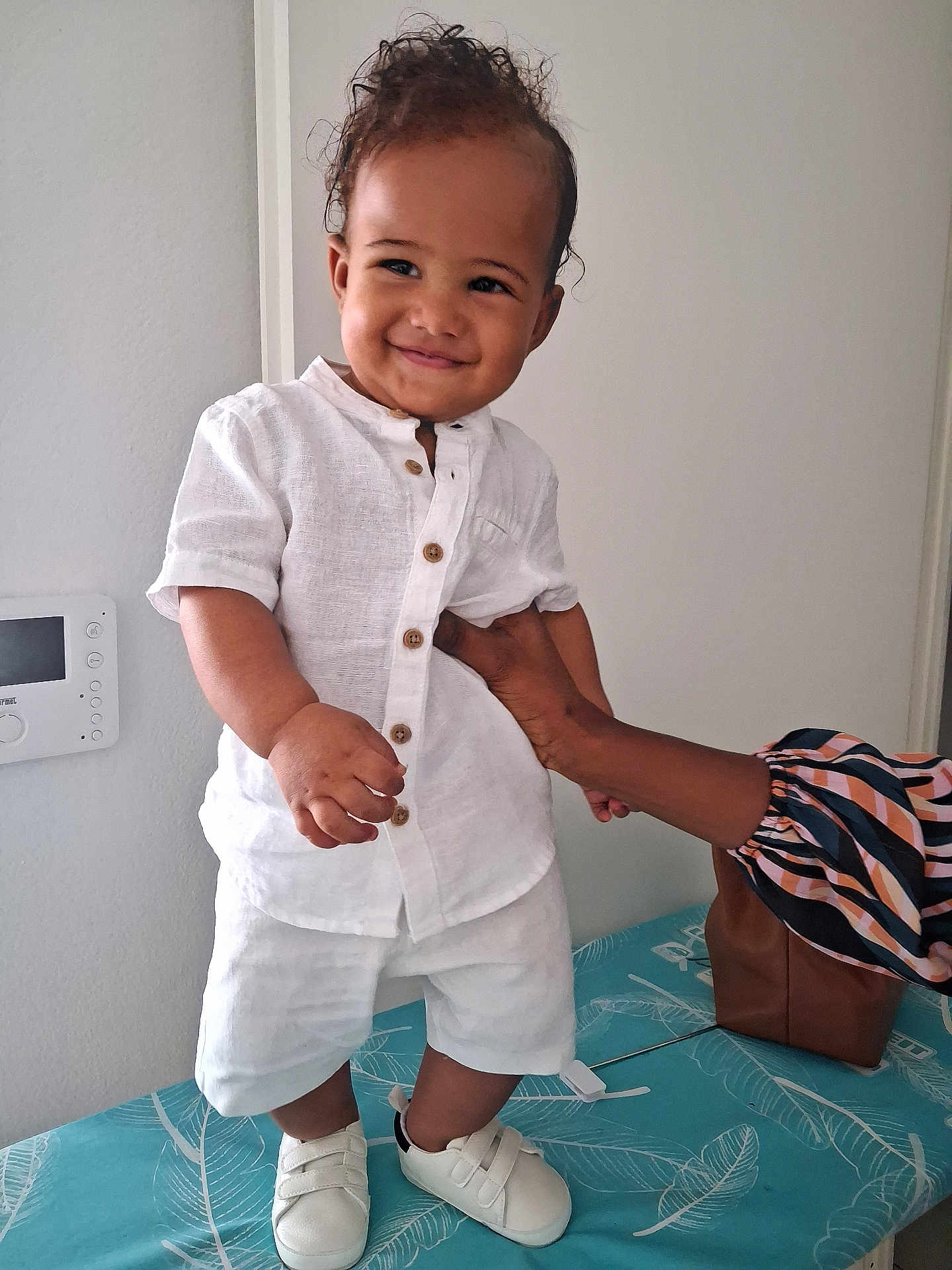 Timaël participe au concours pour gagner de l'argent avec cette photo : toddler, child, smile, white_clothing, shoes, indoor, person, hand, curly_hair, standing, support, joy, patterned_surface, brown_skin, shorts, buttoned_shirt, cute, happy, young_child, baby