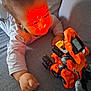 casual, child, closeup, clothing, couch, curious, expression, face, hand, indoor, infant, light_pattern, orange, playtime, plush, relaxed, robot, toddler, toy, young_child