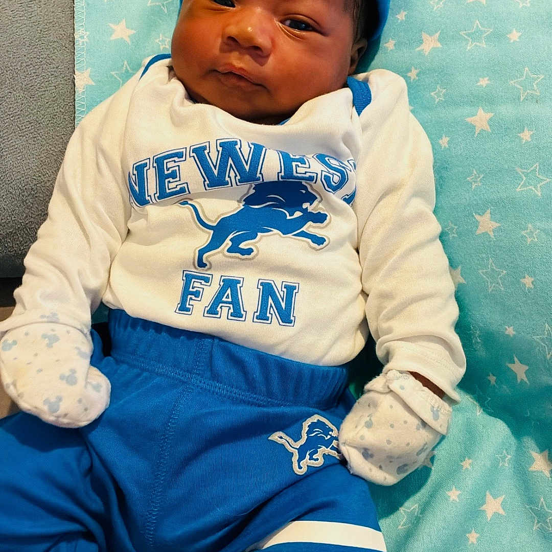 Darion is registered to the contest to win money with this photo: newborn, baby, infant, child, portrait, lying_down, football_fan, sportswear, blue_clothing, white_clothing, cap, mittens, socks, cushion, blanket, stars, indoor, smiling, face, photography