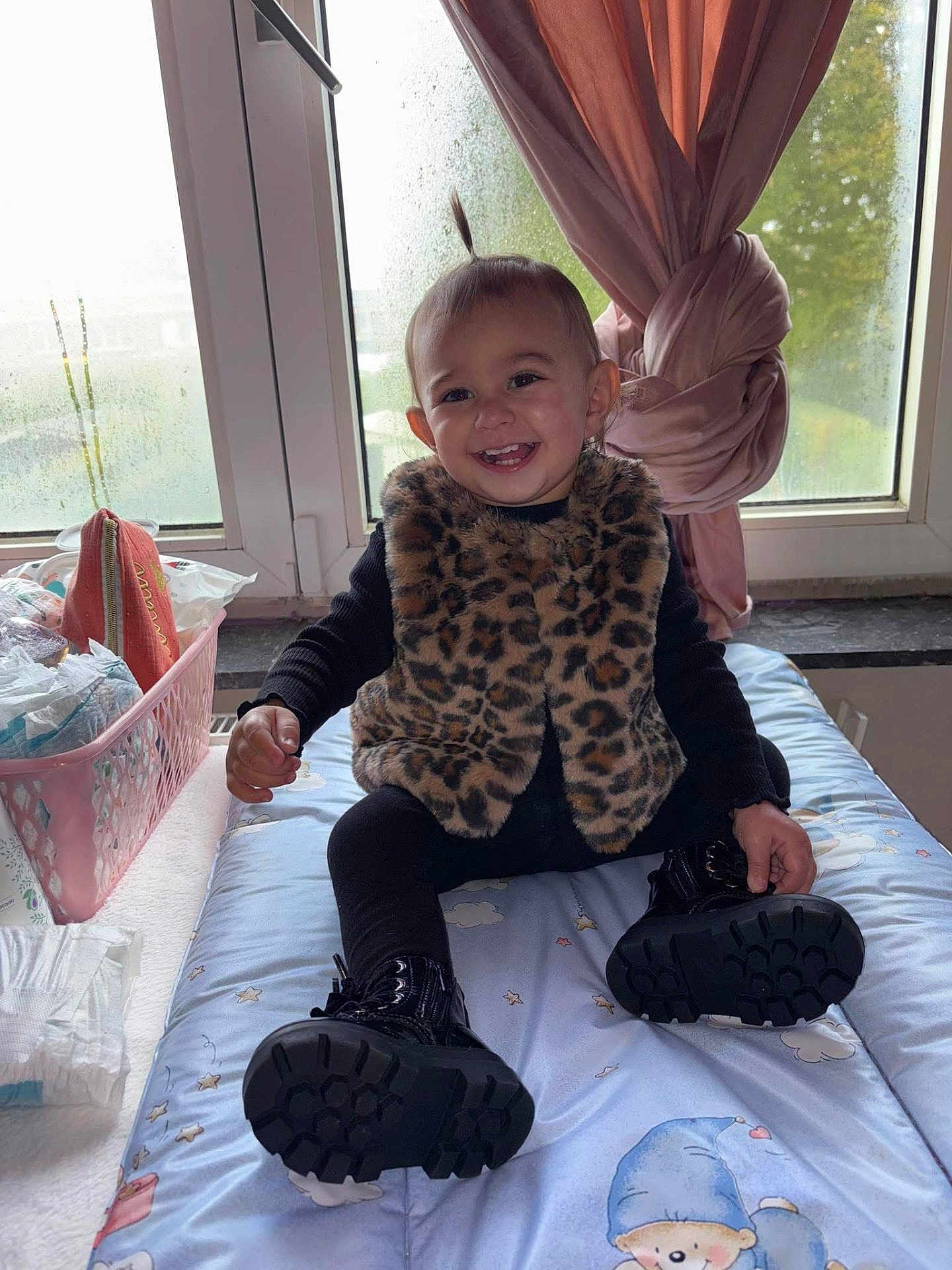 Elyna a rejoint le concours — aidez-le/la à gagner de superbes lots ! toddler, child, smile, leopard_print, vest, black_boots, changing_pad, window, pink_curtains, baby_supplies, diapers, basket, indoor, happy, person, clothing, face, hair, furniture, room