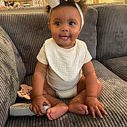 Noelani is registered to the contest to win money with this photo: baby, child, infant, bow, headband, remote_control, toy, couch, sofa, home_interior, living_room, clothing, smile, tongue_out, sitting, cute, plush, furniture, person, happy