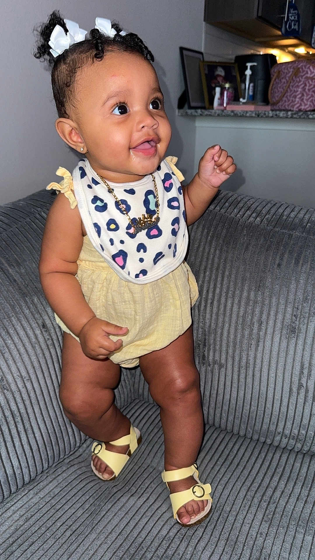 Noelani joined the competition — help win amazing prizes! baby, child, smiling, pigtails, white_bows, yellow_outfit, bib, leopard_print, sandals, couch, indoor, happy, cute, person, portrait, infant, fashion, accessory, jewelry, home