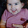baby, child, infant, smiling, happy, portrait, curly_hair, hair_bows, pink_clothing, indoor, seated, being_held, caregiver_hand, face, eyes, teeth, joyful, wooden_table, close_up, photograph