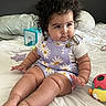 Kianna is registered to the contest to win money with this photo: baby, child, curly_hair, daisy_outfit, purple_clothing, sitting, bed, bedding, pillow, toy, colorful_toy, starfish_toy, indoors, bedroom, portrait, face, smiling, barefoot, infant, photography