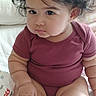 baby, child, curly_hair, portrait, sitting, bed, bedroom, indoor, infant, maroon_clothing, onesie, adorable, serious_expression, round_face, dark_hair, bedding, candid, close_up, person, safe