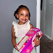 Harmony is registered to the contest to win money with this photo: child, girl, smile, braids, beads, white_top, green_skirt, cake_box, indoor, carpet, wall, doorway, holding, happy, portrait, person, cute, colorful, standing, celebration