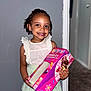child, girl, smile, braids, beads, white_top, green_skirt, cake_box, indoor, carpet, wall, doorway, holding, happy, portrait, person, cute, colorful, standing, celebration