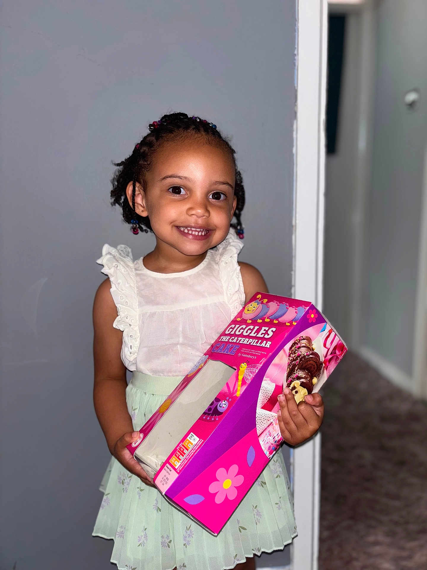 Harmony is registered to the contest to win money with this photo: child, girl, smile, braids, beads, white_top, green_skirt, cake_box, indoor, carpet, wall, doorway, holding, happy, portrait, person, cute, colorful, standing, celebration