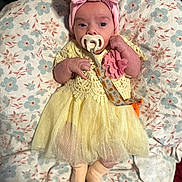 Zara is registered to the contest to win money with this photo: baby, infant, pacifier, headband, bow, dress, yellow_dress, pink, flower_pattern, cushion, blanket, lying_down, cute, small_hands, clothing, newborn, indoors, soft_texture, sleepwear, child