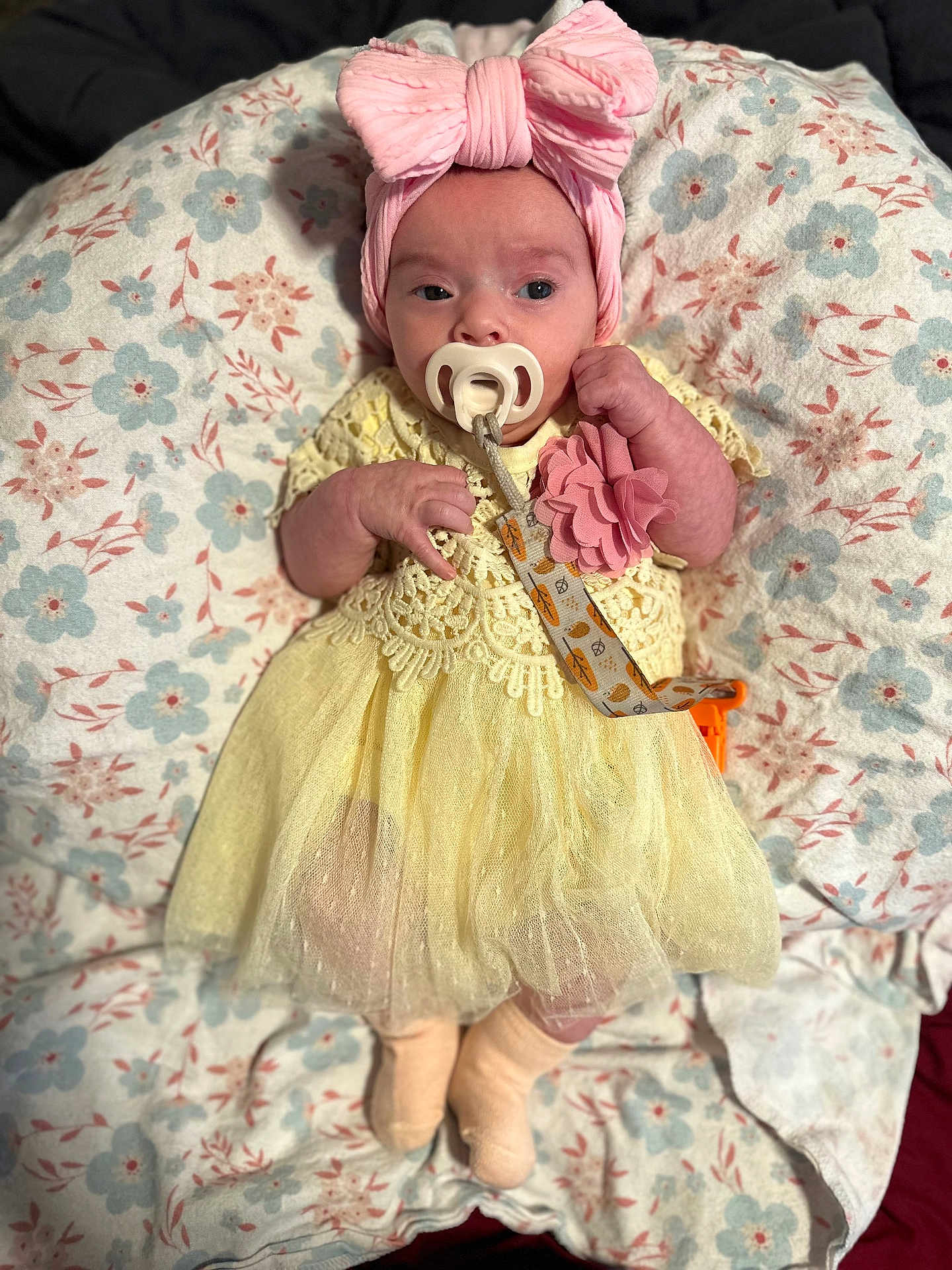 Zara is registered to the contest to win money with this photo: baby, infant, pacifier, headband, bow, dress, yellow_dress, pink, flower_pattern, cushion, blanket, lying_down, cute, small_hands, clothing, newborn, indoors, soft_texture, sleepwear, child