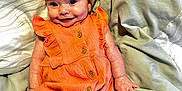 Zara is registered to the contest to win money with this photo: baby, child, infant, smile, orange_outfit, headband, buttoned_clothing, bed, blanket, soft_fabric, cuteness, portrait, happy, person, skin, hand, lying_down, indoors, cozy, young